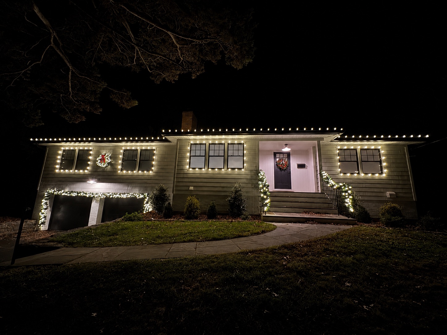 Upscale home at night with warm-white Christmas lights along the roofline and a Tiger Clean truck in the driveway